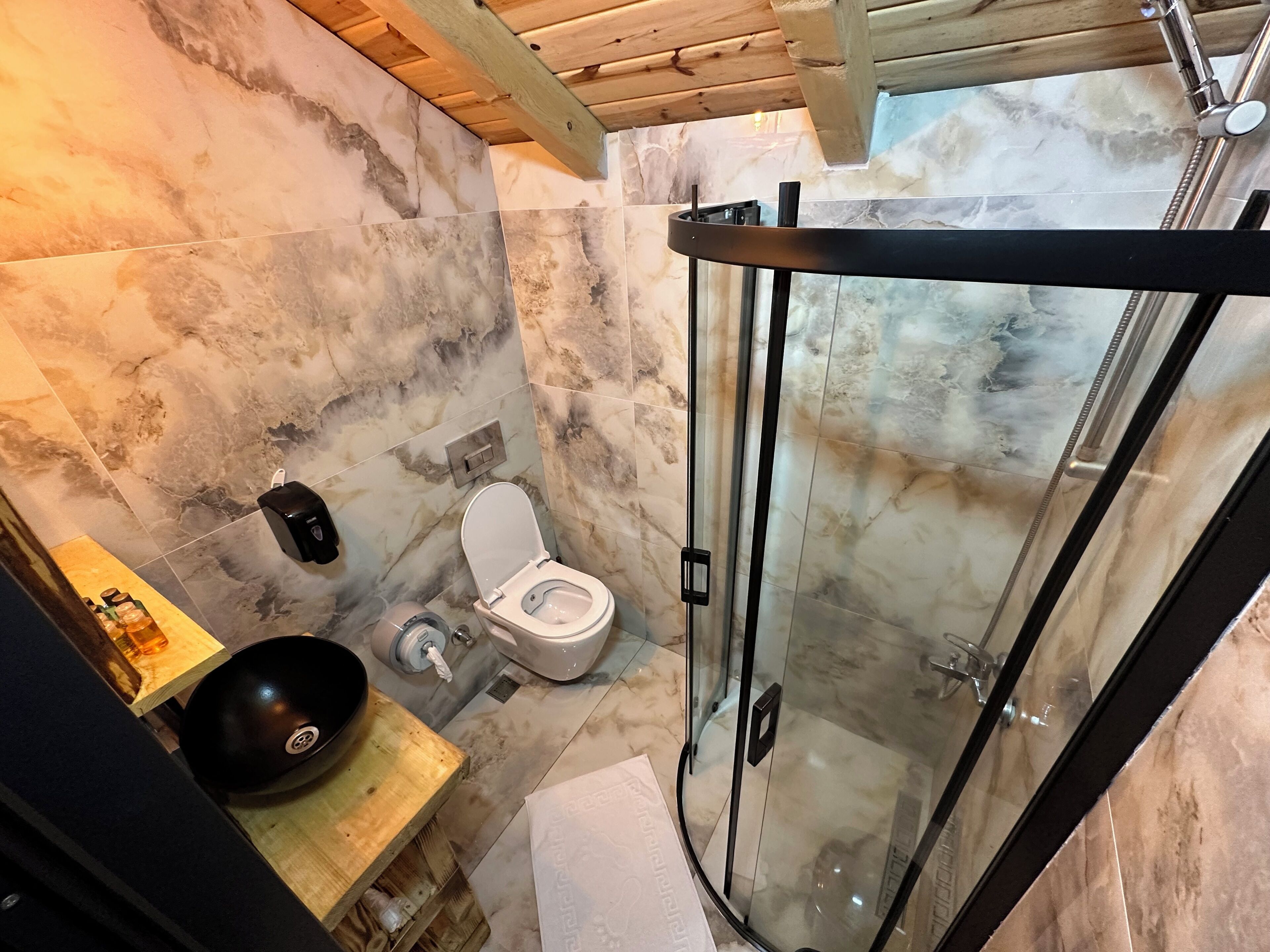 Une salle de bain avec un mur en marbre, des toilettes, un lavabo doté d’un receveur noir et une douche à parois en verre.