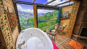 Een kamer met een bubbelbad, houten wanden, uitzicht op groene heuvels en een stenen openhaard.