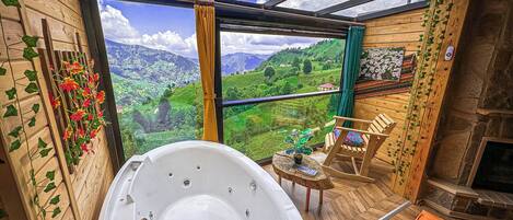 Et værelse med jacuzzi, trævægge, udsigt til grønne bakker og en stenspir.