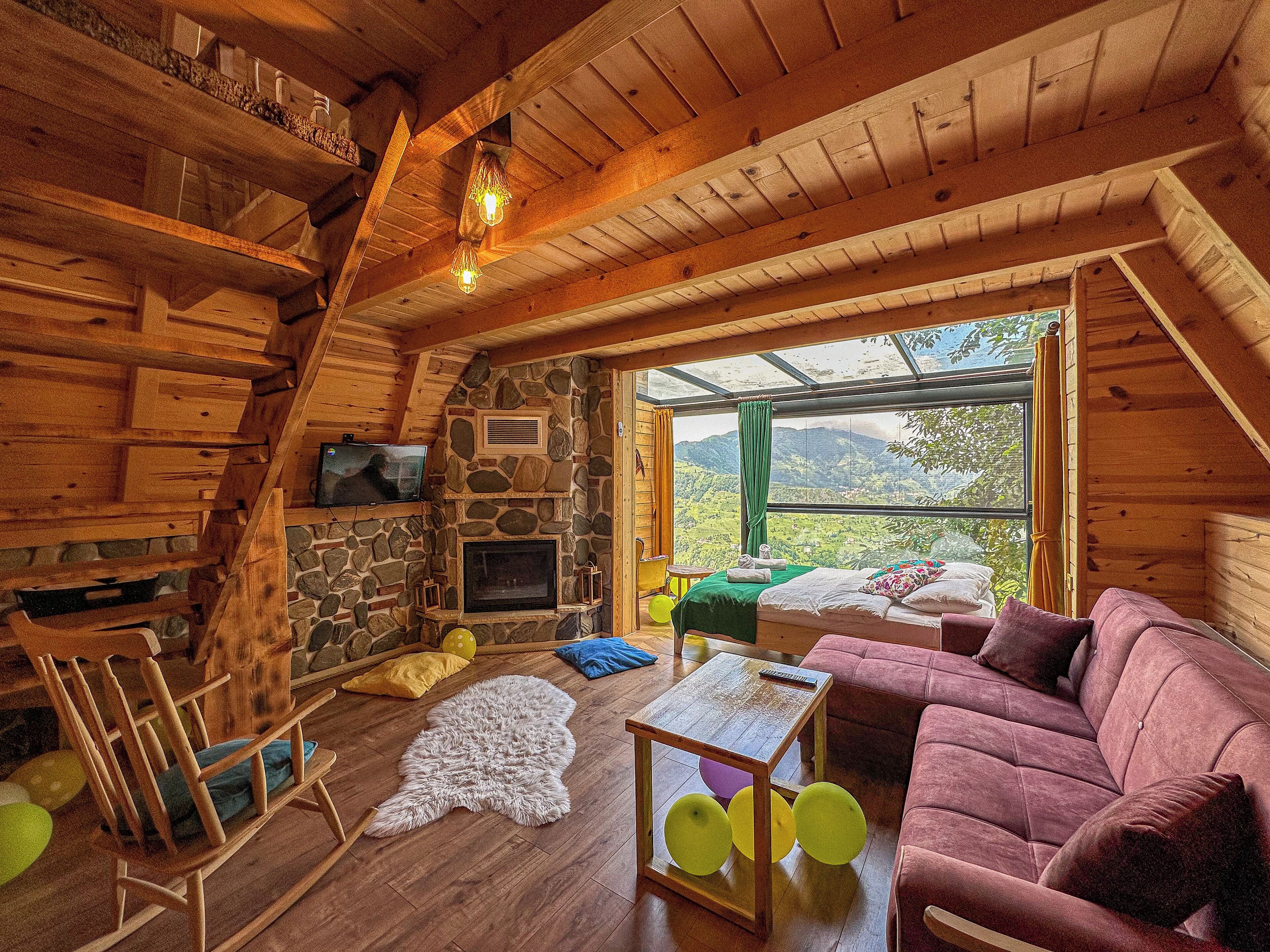 A cozy cabin interior with a stone fireplace, wooden walls, a comfortable couch, and a large window offering a scenic view.