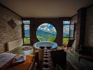 Een kamer met een rond raam met uitzicht op een berg, een badkuip, een houten bank en een bank.