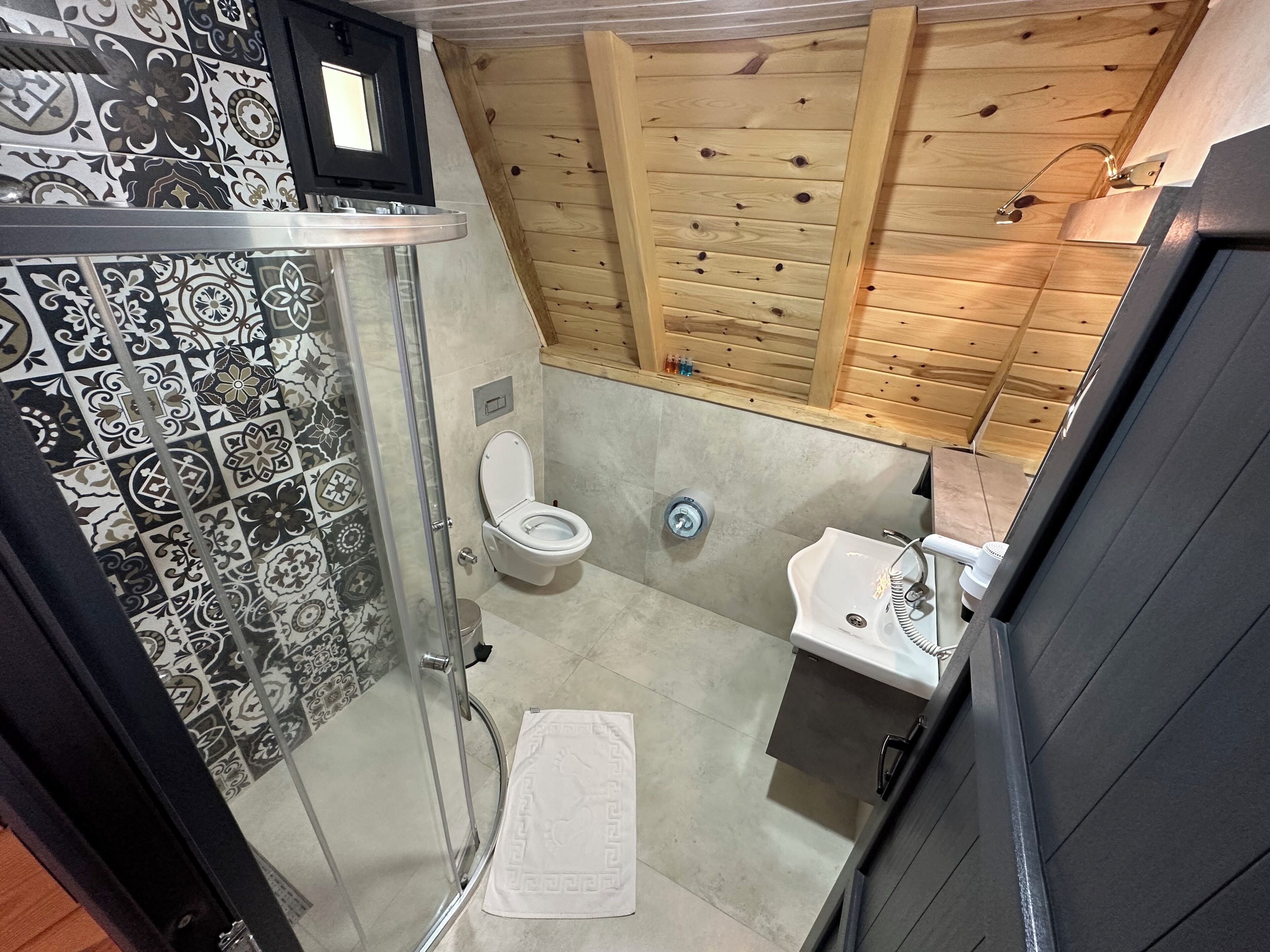 Een badkamer met douche, toilet en wastafel, met tegelwerk met een patroon en houten lambrisering.