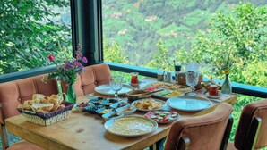 Een eettafel gedekt met verschillende gerechten, een vaas met bloemen en uitzicht op een bergachtig landschap door een raam.