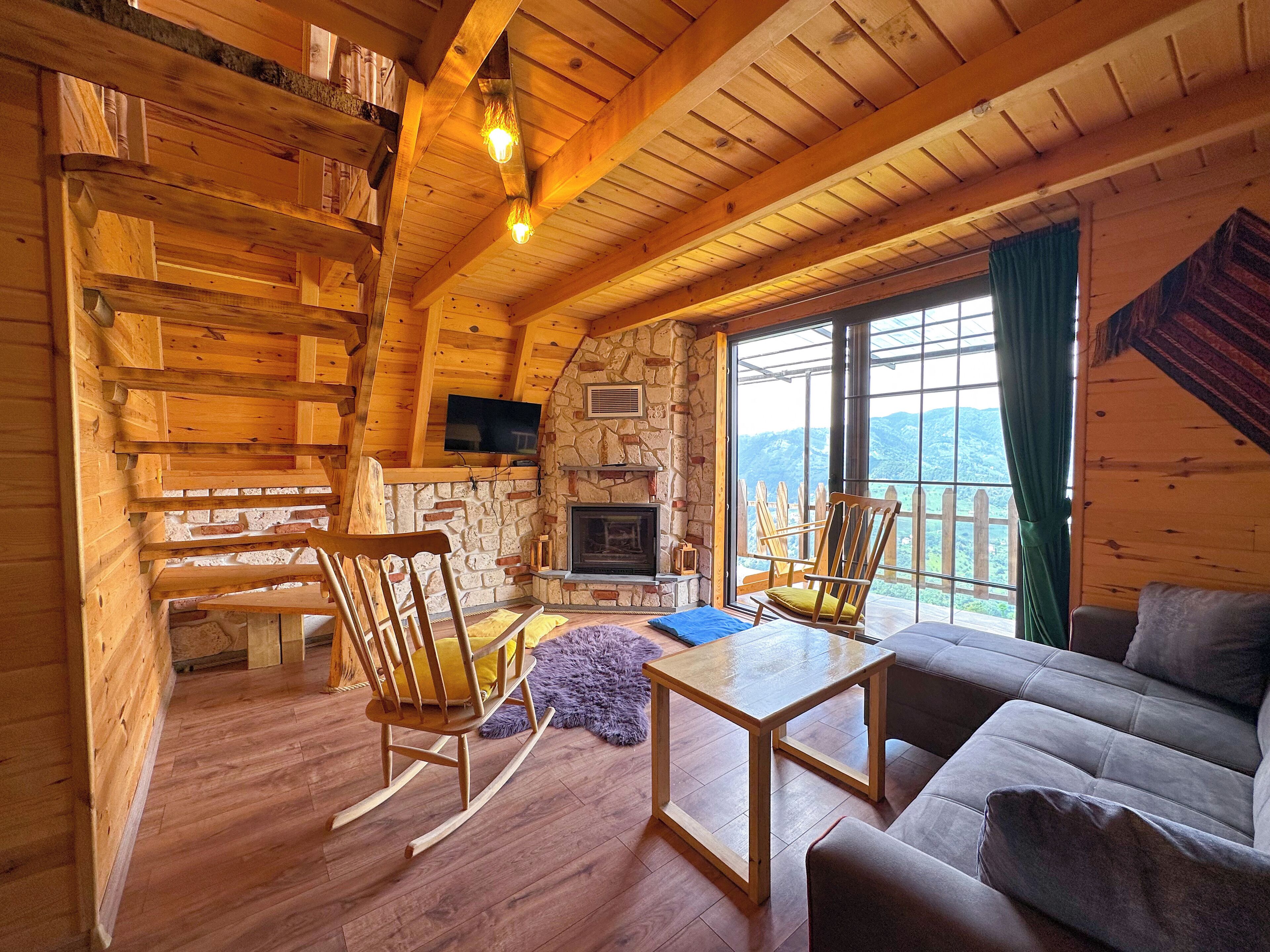 A cozy cabin interior with a stone fireplace, wooden furniture, and a large window offering a scenic view.