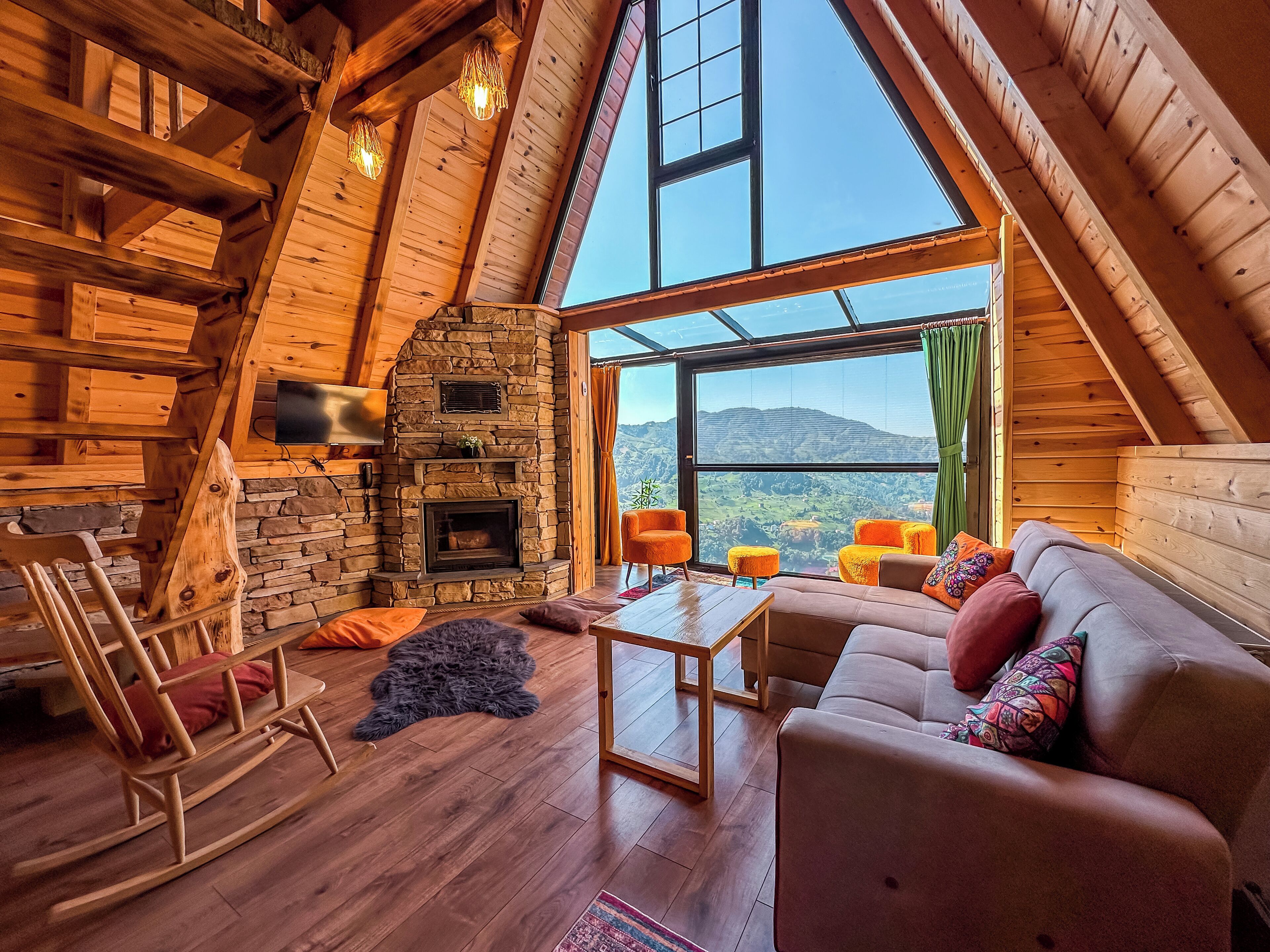 Un accogliente interno di una baita in legno con camino in pietra, travi in legno, un'ampia finestra con vista sulle montagne e una zona salotto confortevole.