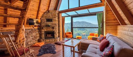 Interior acogedor de una cabaña de troncos con chimenea de piedra, vigas de madera, un ventanal con vistas a la montaña y una zona de estar confortable.