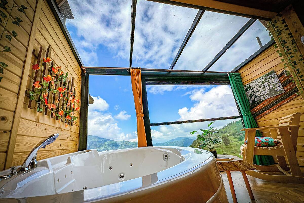 A wooden cabin interior with a large window, a hot tub, and a wooden chair.