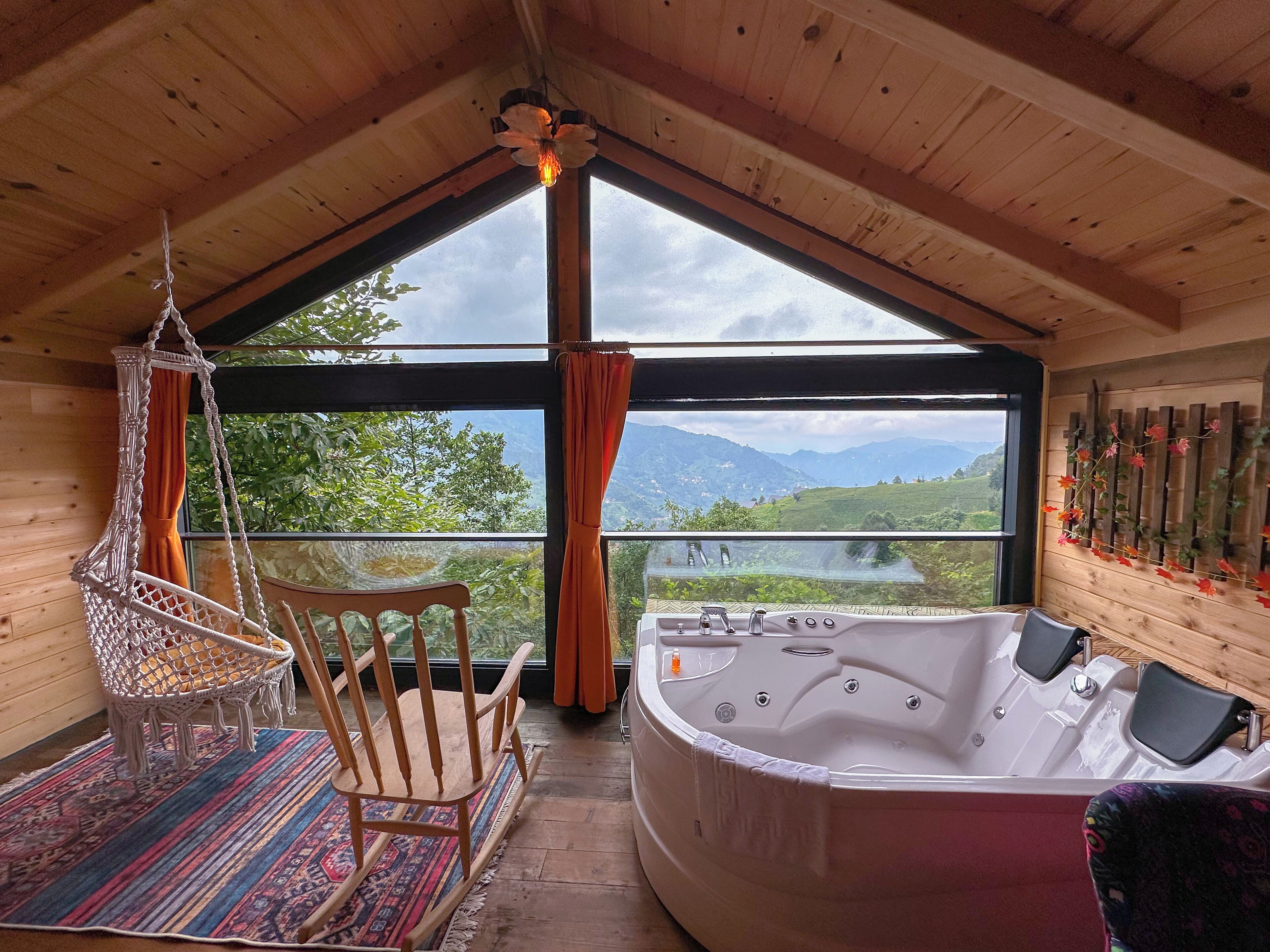 A wooden cabin with a large window, a hot tub, a macramé chair, and a patterned rug.
