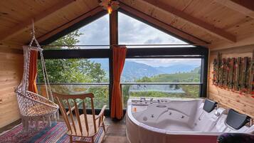 A wooden cabin with a large window, a hot tub, a macramé chair, and a patterned rug.