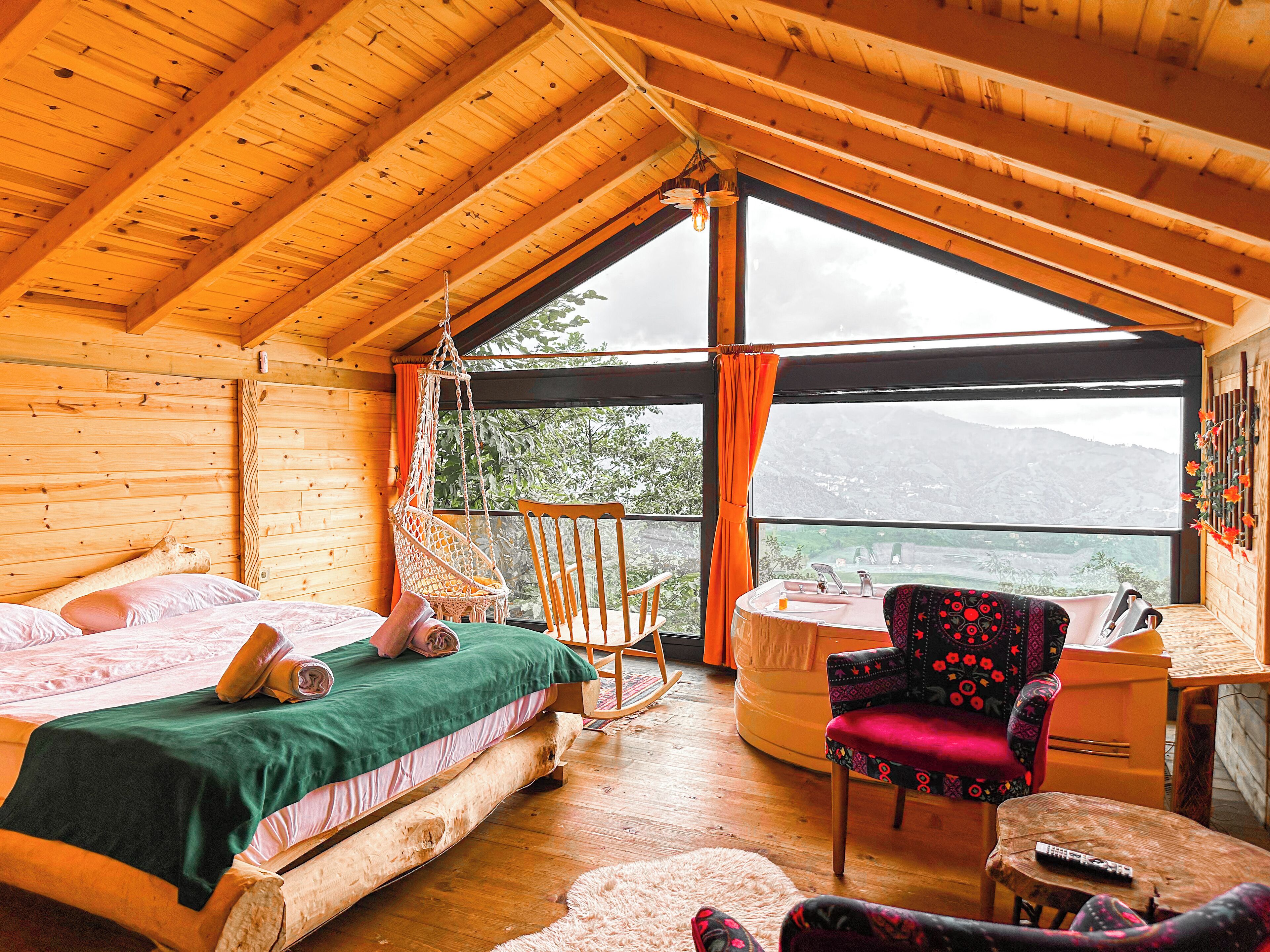 A cozy wooden cabin room with a bed, a rocking chair, a desk, and a view of the outdoors.