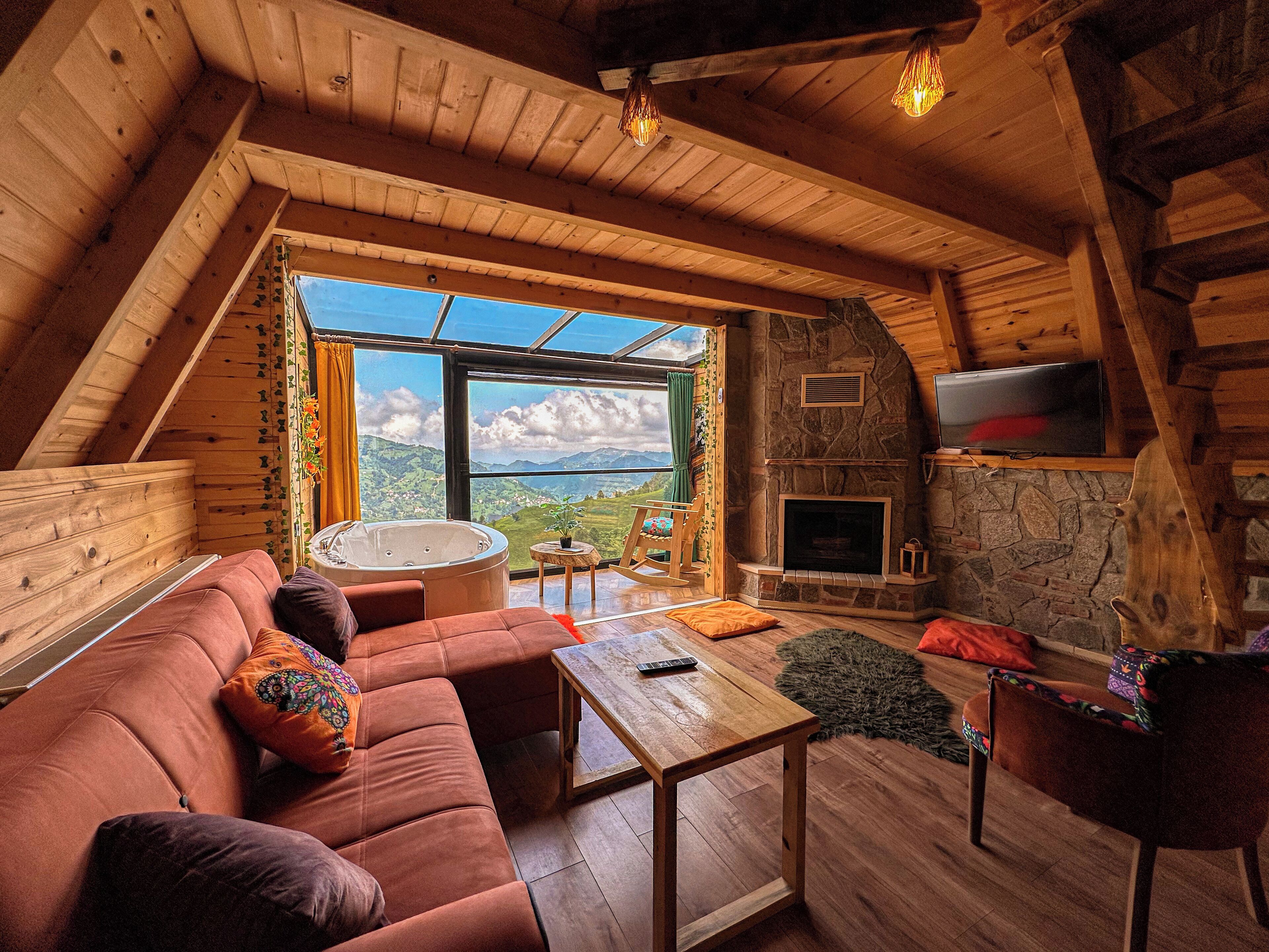 Ein gemütliches Blockhaus-Interieur mit Kamin aus Stein, Holzbalkendecke und großem Fenster mit Blick auf eine gebirgige Landschaft.