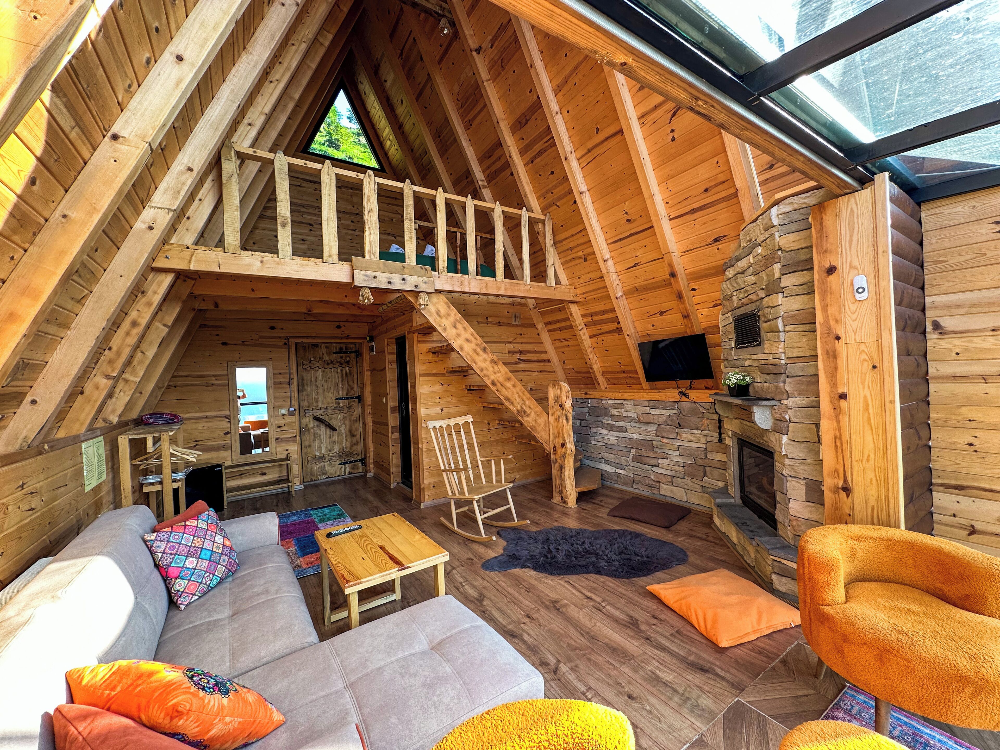 A cozy loft-style room with a wooden ceiling, a stone fireplace, a large window, and a sofa with colorful cushions.