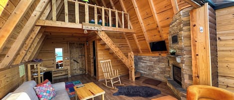 A cozy loft-style room with a wooden ceiling, a stone fireplace, a large window, and a sofa with colorful cushions.