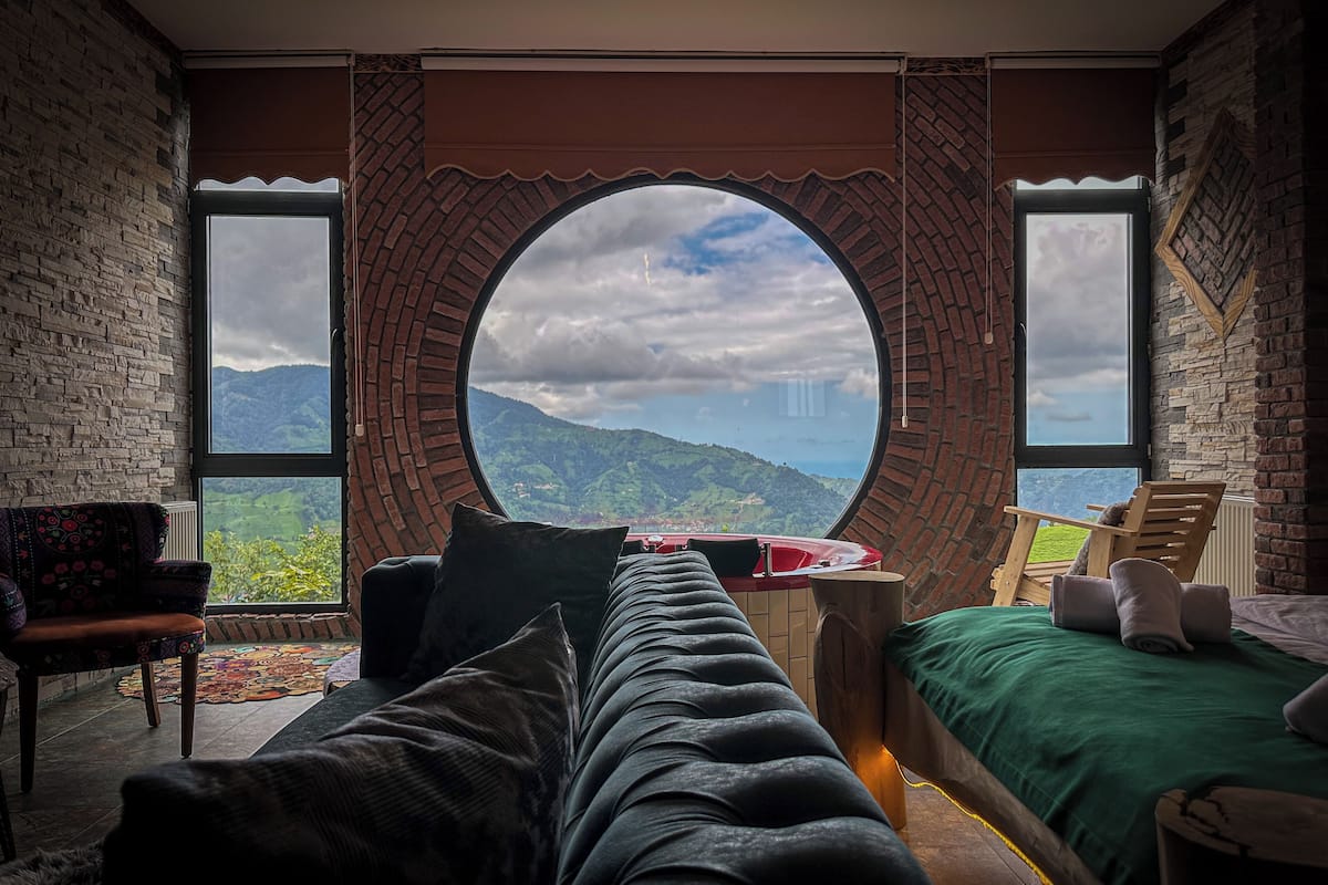 A room with a large circular window offering a mountain view, a sofa, a chair, and a small table with a red object on it.