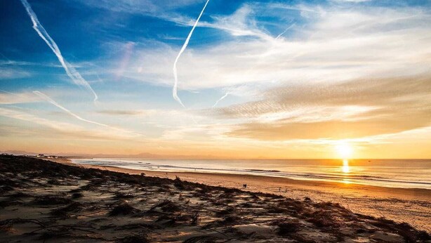 Ein Strand mit Sonnenuntergang, klarem Himmel und sichtbaren Kondensstreifen.