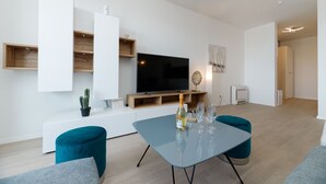 Ein modernes Wohnzimmer mit Flachbildfernseher, Holzregalen, einem Glastisch mit Weingläsern und Türkis-farbenen Stühlen.