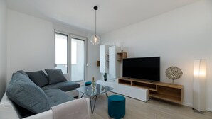 Ein modernes Wohnzimmer mit grauem Sofa, einem Glastisch, einem Flachbildfernseher auf einem Holzboden und einem großen Fenster, das für natürliches Licht sorgt.