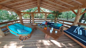 A wooden deck with a covered seating area featuring a rocking chair, a sofa, and a table with a fruit bowl and glasses. - Chalet à Proximité de L'océan Avec Piscine Insolite (Queyrac)