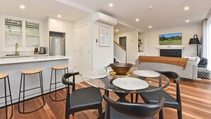 A modern kitchen with a built-in refrigerator, a dining area with a glass table and black chairs, and a living room with a sofa and a television.