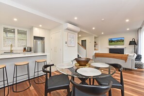 Una cocina moderna con refrigerador empotrado, un comedor con mesa de vidrio y sillas negras, y una sala de estar con sofá y televisor.