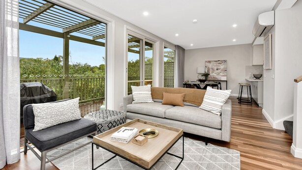 A modern living room with a grey sofa, a black armchair, and a coffee table. There is a balcony with a view of greenery.