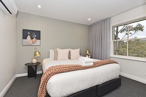 Un dormitorio con cama, mesitas de noche, una ventana con vistas y un cuadro en la pared.