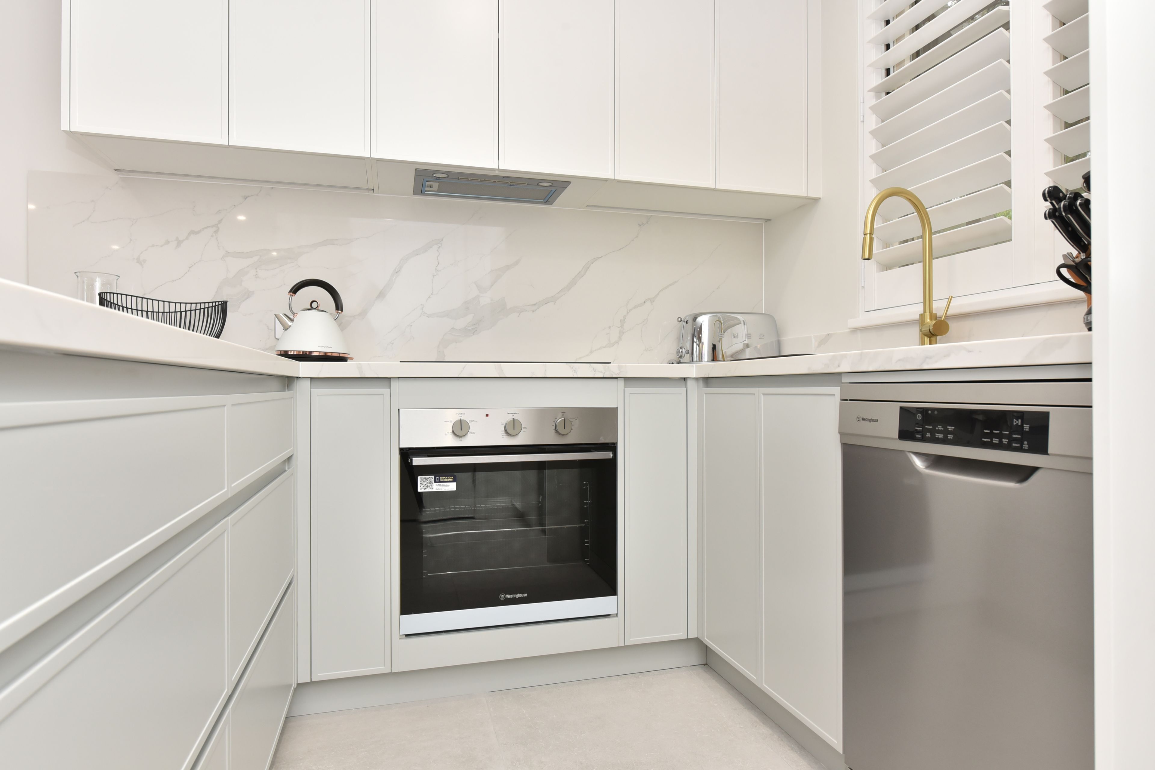 A modern kitchen with white cabinetry, a built-in oven, a dishwasher, and a kettle on the countertop.