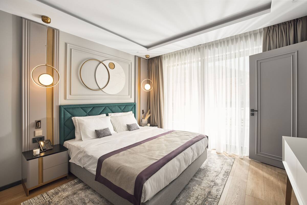 Ein modernes Hotelzimmer mit einem großen Bett, einem Nachttisch mit Lampe, einem an der Wand befestigten Spiegel und einer Tür rechts.