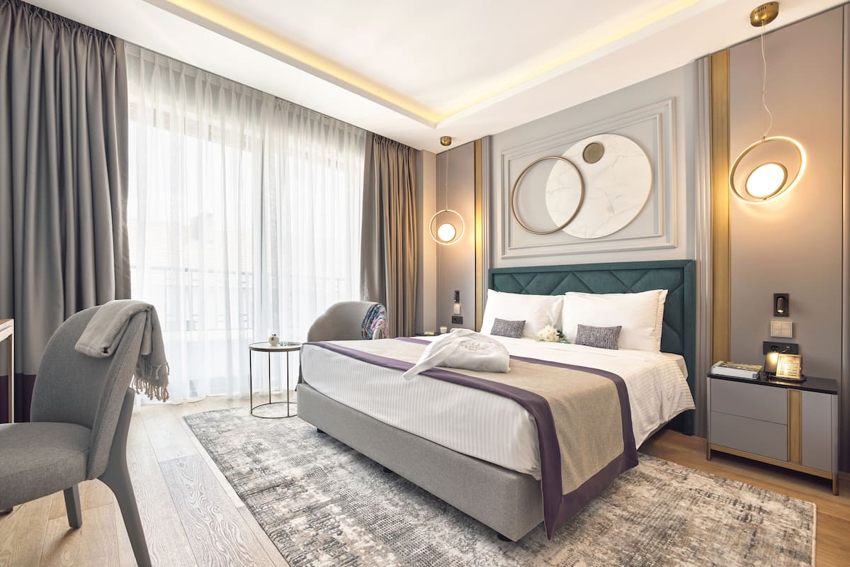 Ein modernes Hotelzimmer mit einem großen Bett, einem Nachttisch, einem Sessel und Blick auf die Stadt durch große Fenster.