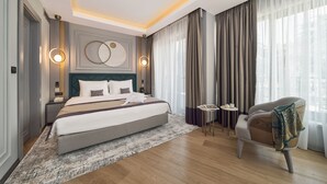 Een moderne hotelkamer met een groot bed, een zithoek met een fauteuil, een tafeltje en uitzicht op de omgeving door dunne gordijnen.