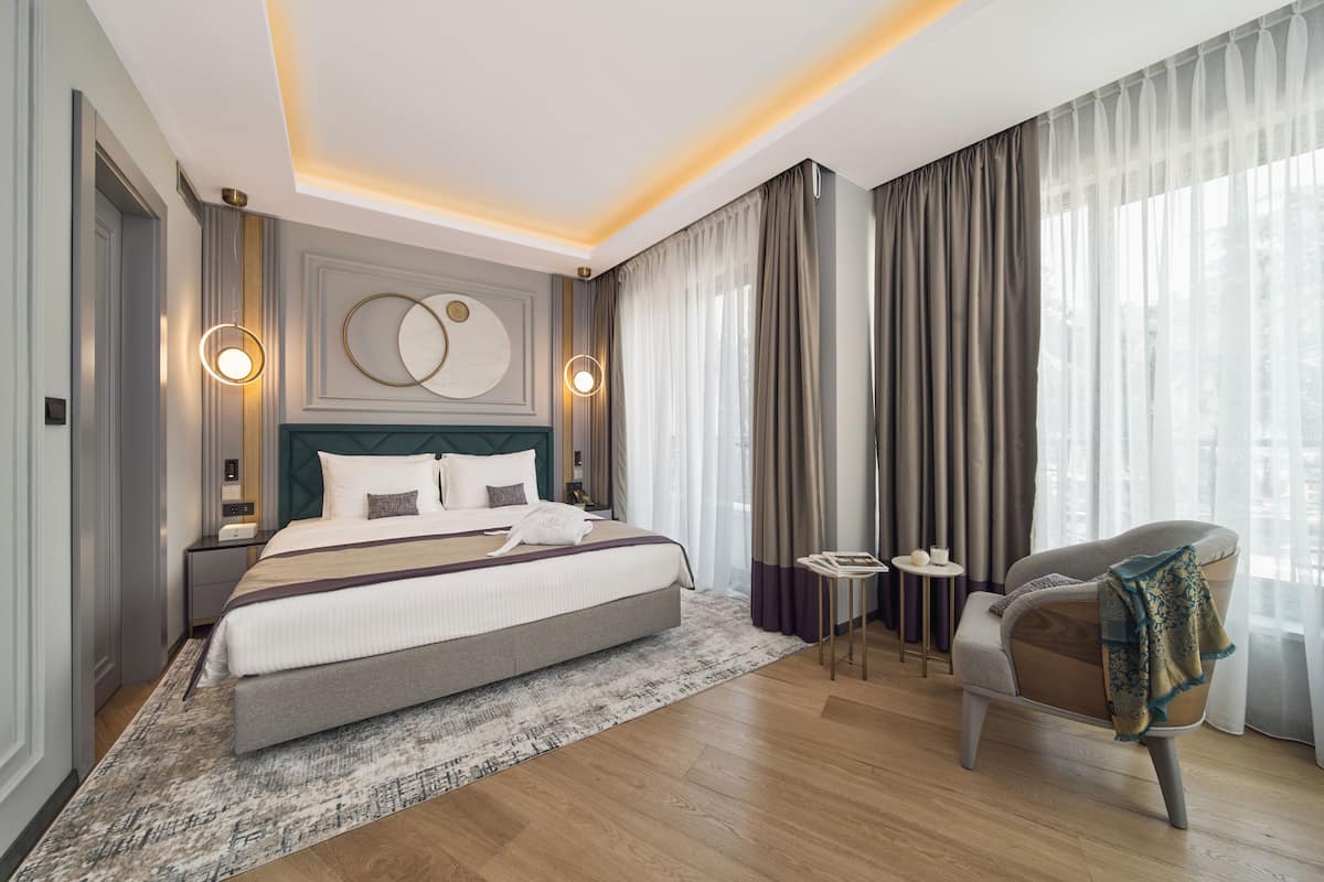Ein modernes Hotelzimmer mit einem großen Bett, einer Sitzecke mit Stuhl und Tisch sowie Blick ins Freie durch transparente Vorhänge.