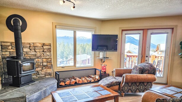 A cozy living room with a stone fireplace, a television, and a view of mountains through large windows. - Pet-Friendly 2-Bedroom Condo Near Hot Springs (Radium Hot Springs)