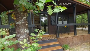 Uma área externa coberta com deck de madeira, mesa e cadeiras, e uma grande árvore com folhas verdes.