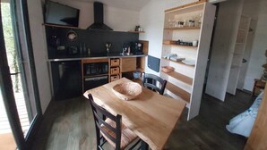 Uma cozinha compacta com uma mesa de madeira, cadeiras e um cesto. Há uma TV fixada na parede e um relógio sobre a porta.