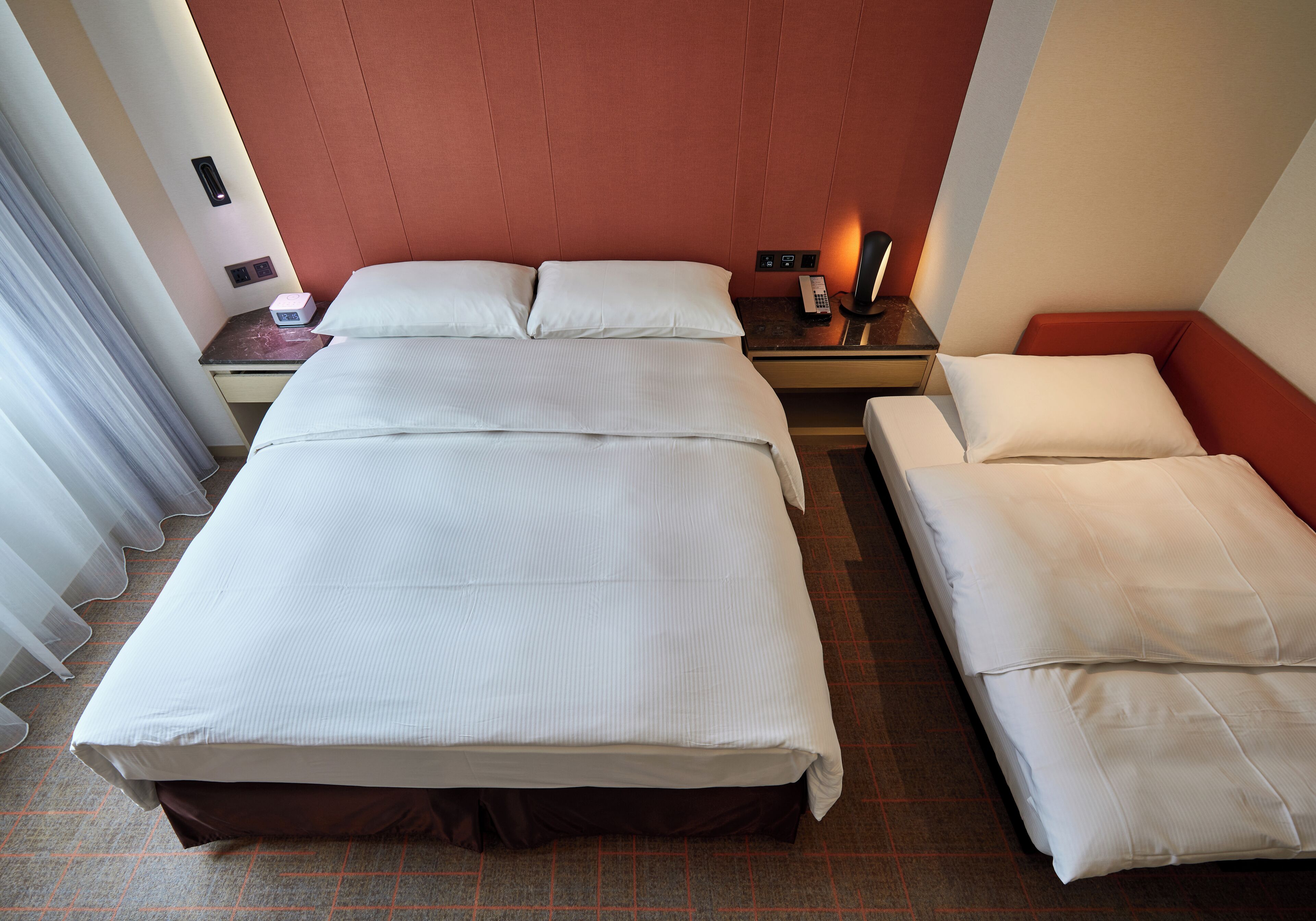 A hotel room with a large bed, a smaller bed, a nightstand with a lamp, and a window with curtains.