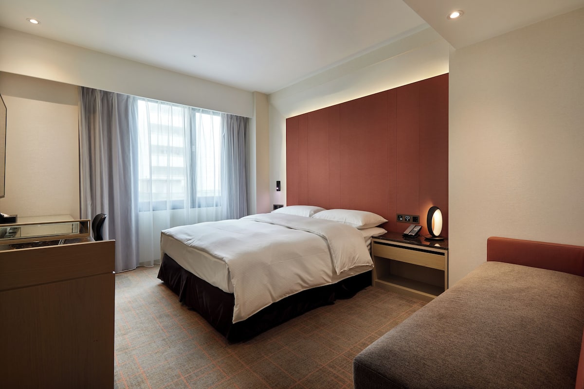 A hotel room with a bed, a desk, a chair, a desk lamp, and a window with curtains.