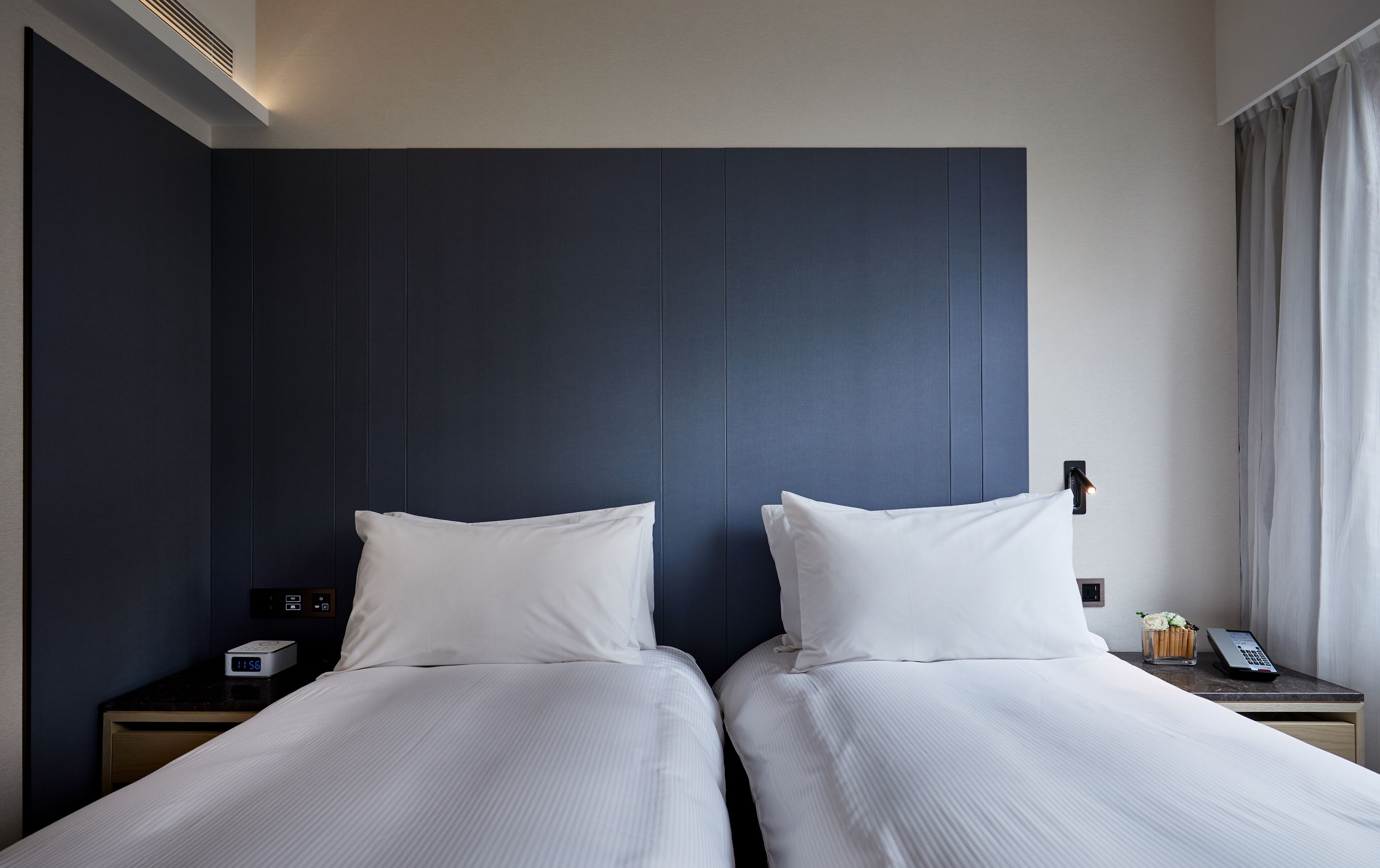 A hotel room with two beds, a nightstand with a phone and a digital clock, and a window with curtains.
