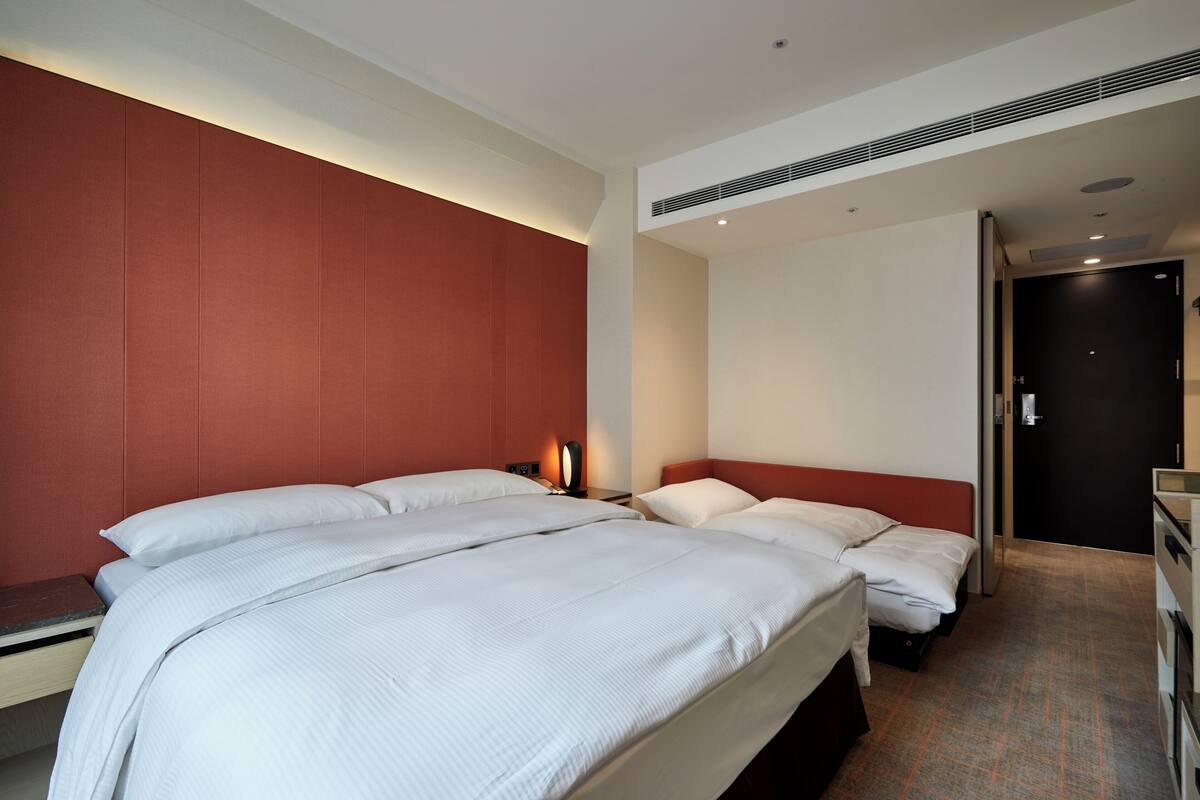 A hotel room with two beds, a red accent wall, a bedside table with a lamp, and a door leading to another room.
