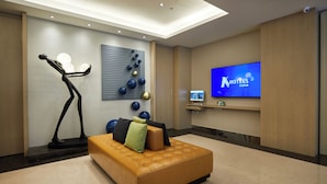 Un hall d’hôtel moderne doté d’un grand écran de télévision affichant le nom de l’hôtel, d’un espace détente avec un canapé et des coussins, d’œuvres d’art décoratives murales et d’un tapis de course.