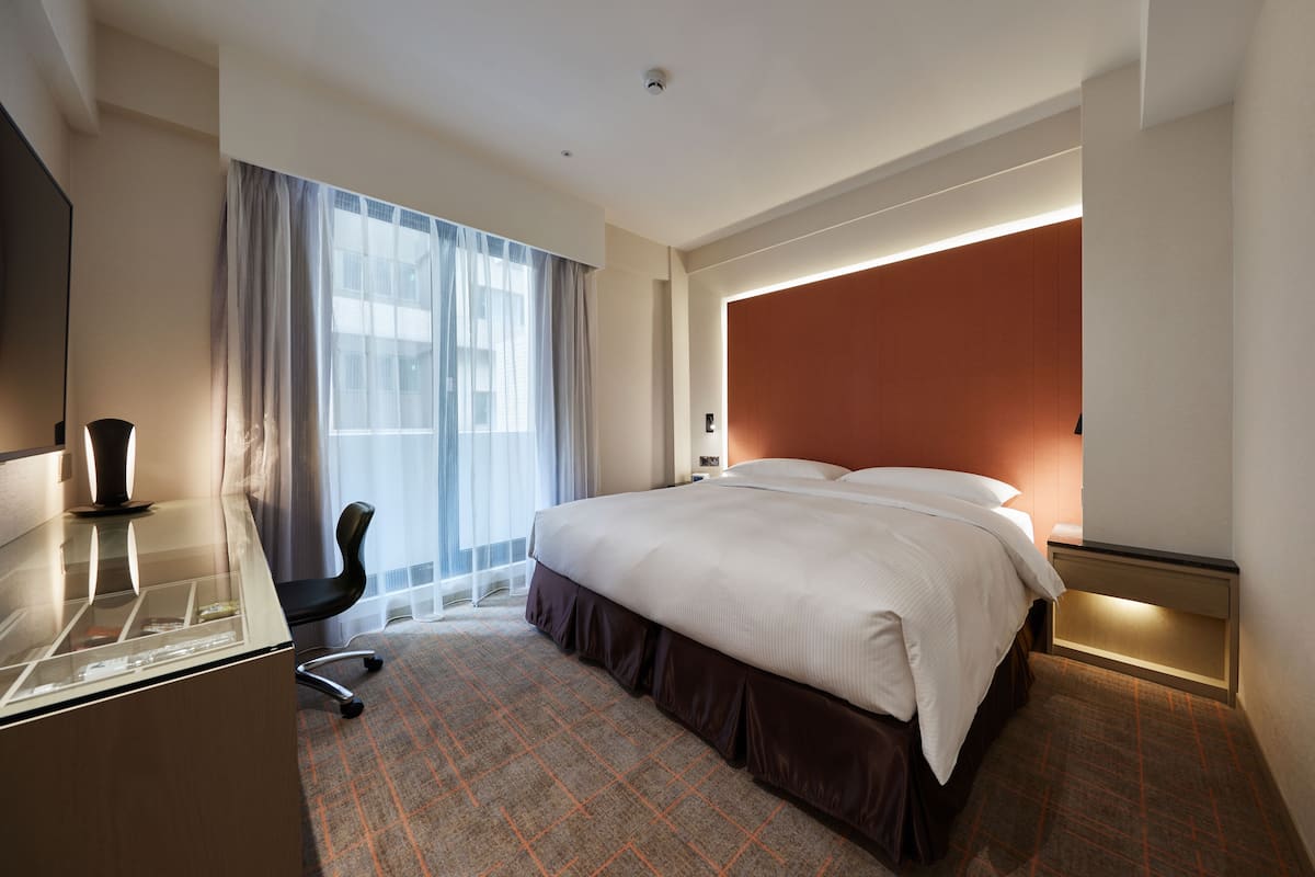 A hotel room with a bed, a desk, a chair, a window with curtains, and a wall-mounted light.