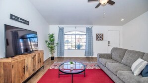 A living room with a flat-screen TV, a ceiling fan, a grey sofa, a red carpet, and a large window with curtains. - LUXURY Living 2 (Yuma)