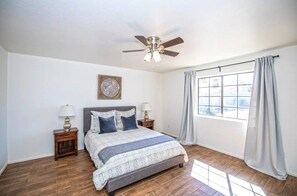 A bedroom with a bed, two bedside tables, a ceiling fan, a window with curtains, and a decorative wall piece.