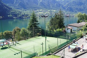 A tennis court with a net, surrounded by a fence, overlooking a lake with a village in the background.