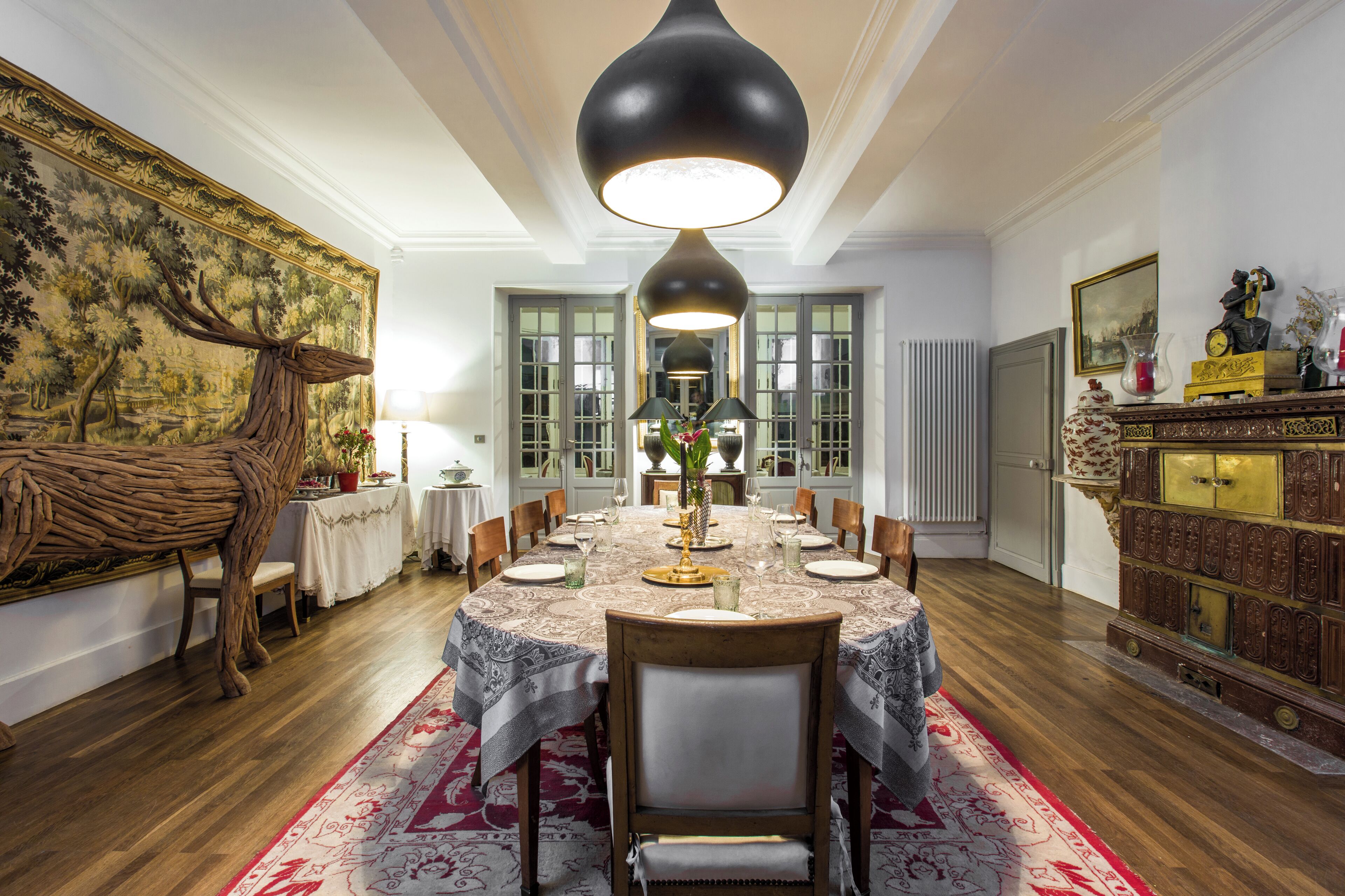 A dining room with a round table set for six, surrounded by chairs. There is a large tapestry on the wall, a wooden cabinet with glass doors, and a radiator on the wall.