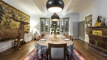 A dining room with a round table set for six, surrounded by chairs. There is a large tapestry on the wall, a wooden cabinet with glass doors, and a radiator on the wall.