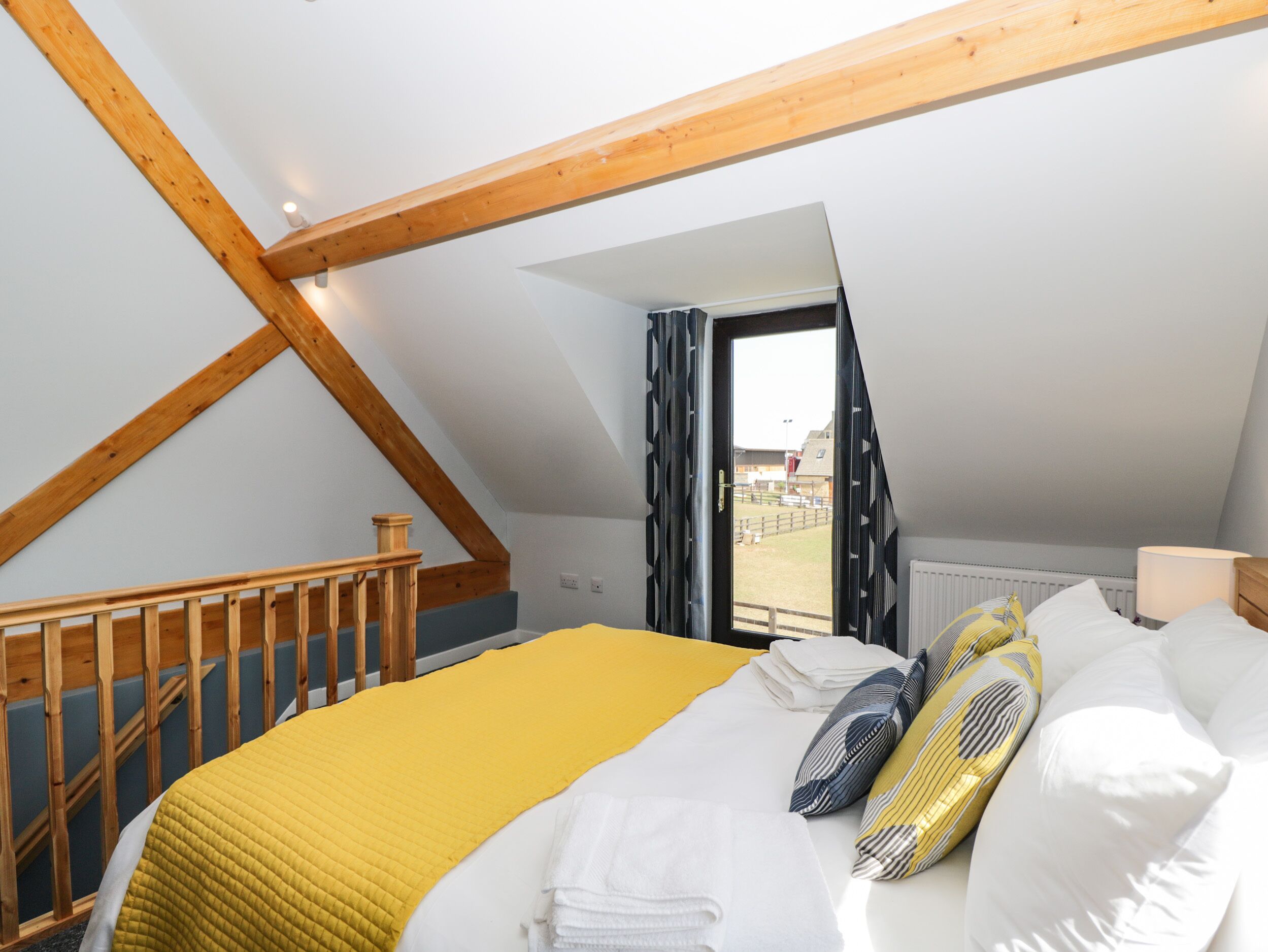 A bedroom with a sloped ceiling, a wooden staircase, a bed with a yellow blanket, and a window with a view of an outdoor area.