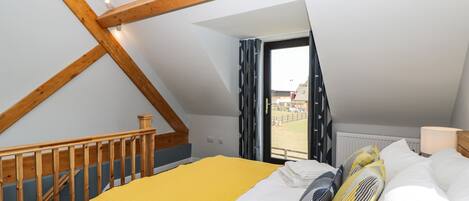 A bedroom with a sloped ceiling, a wooden staircase, a bed with a yellow blanket, and a window with a view of an outdoor area.
