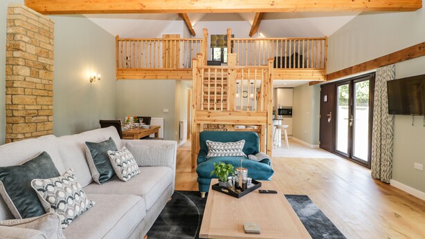 A spacious living area with a high ceiling and exposed wooden beams, a cozy sofa set, a dining table, and a view of the kitchen and outdoor area. - Petals Patch (Cheltenham)