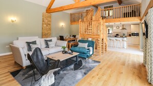 A spacious living area with a high ceiling and exposed wooden beams, a cozy seating area with a sofa and chairs, a dining table, and a kitchen in the background. - Petals Patch (Cheltenham)