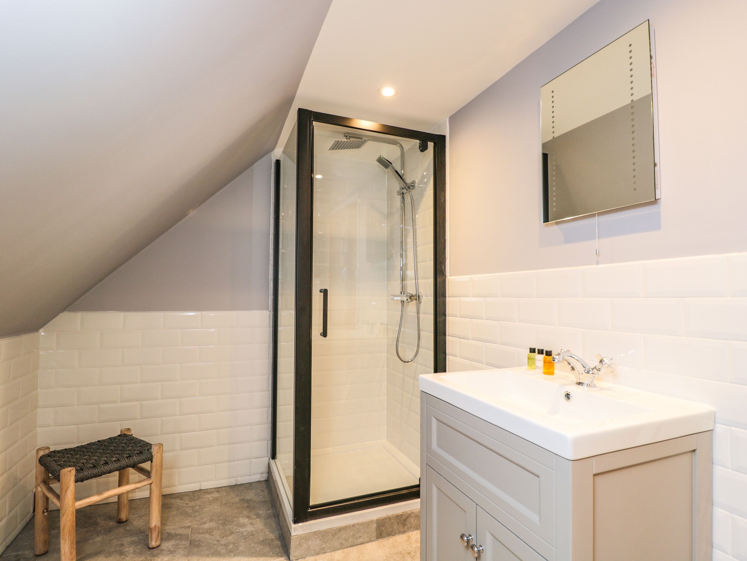 A modern bathroom with a glass shower enclosure, a wooden stool, a white vanity, and a mirror above the sink.